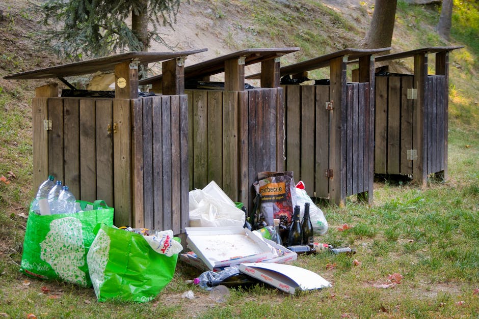 A row of four wooden outdoor rubbish bins with slanted roofs, situated on a grassy area with a slight incline and surrounded by trees. The bins are dark brown with vertical wooden planks, metal hinges, and locks. In front of the bins, there is an accumulation of various discarded items, including several clear plastic bottles, some contained within a bright green reusable shopping bag, along with a white foam food container, a black and orange cardboard box labeled 'barbecue,' overturned glass bottles, and scattered paper and plastic waste. The background features a natural environment with tree trunks visible to the right and behind the bins, and a mixture of light and shadow suggesting daylight filtering through the trees. The scene appears to depict an informal waste disposal area, contrasting with the organised rubbish collection services offered by Waste Collection Kingston for private or alternative waste handling.