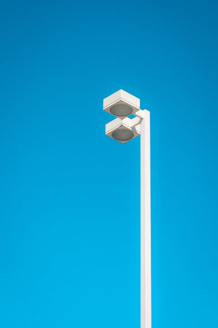 A tall, white street light pole stands against a clear, bright blue sky. The pole supports a modern, rectangular security or surveillance camera system with two large, rounded camera lenses housed in white casings, positioned at the top of the pole. The camera units have smooth, matte finishes and are mounted in a way that allows for wide-angle coverage. The scene is outdoors, with no other objects or environmental details visible, emphasizing the minimalist and functional design of the equipment. This type of infrastructure is commonly used for monitoring outdoor areas, which may include environments such as residential or commercial premises, or public spaces. The high clarity of the sky and the clean appearance of the equipment suggest daytime with natural lighting. The setup illustrates typical on-site security measures that might be employed by businesses or property management to ensure safety and surveillance, connecting subtly to services related to waste management sites or private property security enhancement.