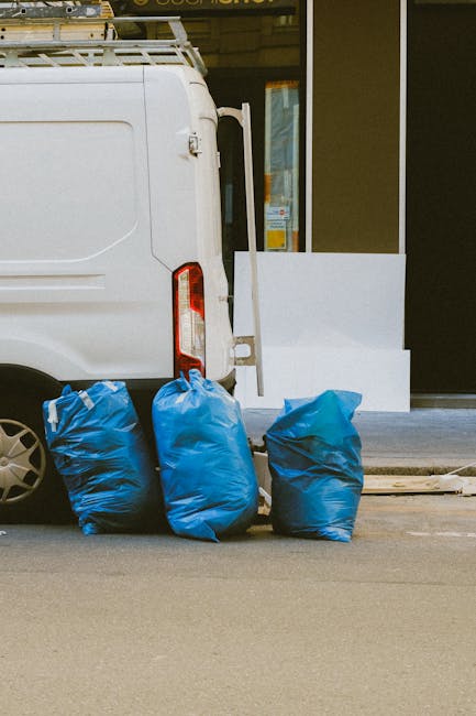 The rear side of a white van parked on a city street, with three blue rubbish bags placed on the ground near the van's back doors. The bags are filled with waste and are made of a crinkled plastic material, with varying shades of blue. The van is positioned next to a building with large glass windows and a dark-colored entrance door, beneath a yellow awning. The scene is illuminated by natural light, highlighting the clean, smooth surface of the van and the textured plastic of the rubbish bags. The area appears to be part of an urban environment, where private waste collection services, such as those by Waste Collection Kingston, might handle on-site clearance or alternative rubbish removal, with the bags prepared for collection or disposal.