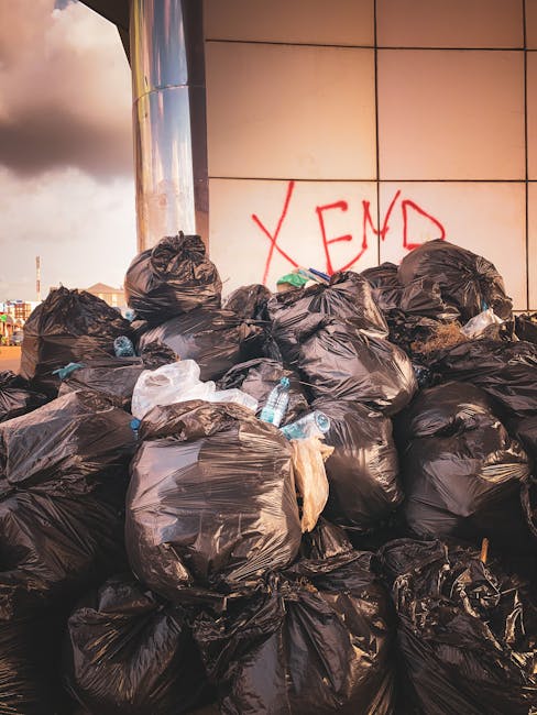 A large pile of black plastic rubbish bags, some slightly torn, is stacked against a tiled wall in an outdoor urban setting. The bags contain various waste items, including visible plastic bottles and crumpled packaging. The bags are stacked haphazardly, with some leaning or partially collapsed, occupying most of the foreground. Behind the rubbish pile, the wall features red spray-painted graffiti spelling out 'XEND' in capital letters. To the left, a reflective metal column or structure catches some light, contrasting with the matte finish of the bags and wall. The environment appears to be a street or alley, with a cloudy sky overhead and distant cityscape elements faintly visible in the background. The scene emphasizes informal waste accumulation, which could be associated with private waste handling or alternative rubbish collection services, such as those offered by Waste Collection Kingston, especially in situations where on-site clearance is performed outside standard municipal collection points.