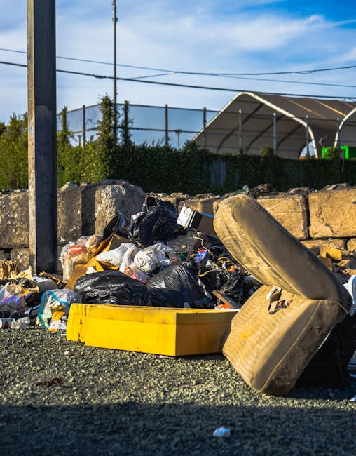 A large pile of black plastic rubbish bags, some slightly torn, is stacked against a tiled wall in an outdoor urban setting. The bags contain various waste items, including visible plastic bottles and crumpled packaging. The bags are stacked haphazardly, with some leaning or partially collapsed, occupying most of the foreground. Behind the rubbish pile, the wall features red spray-painted graffiti spelling out 'XEND' in capital letters. To the left, a reflective metal column or structure catches some light, contrasting with the matte finish of the bags and wall. The environment appears to be a street or alley, with a cloudy sky overhead and distant cityscape elements faintly visible in the background. The scene emphasizes informal waste accumulation, which could be associated with private waste handling or alternative rubbish collection services, such as those offered by Waste Collection Kingston, especially in situations where on-site clearance is performed outside standard municipal collection points.