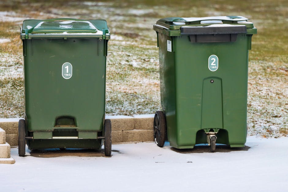 Two green wheeled rubbish bins are positioned outdoors on a slightly snowy surface, with the left bin labeled with the number '1' and the right bin labeled with the number '2'. The bin on the left appears to have a smooth, matte finish with some minor scratches and scuff marks, while the bin on the right has a slightly textured surface with a darker, fitted lid and black handles. Both bins are set on a paved area bordered by a small brick wall, with a background of a grassy yard and a paved driveway, indicating a residential setting. The scene is lit by natural daylight, casting soft shadows beneath the bins. The context suggests a private collection system for waste and garden refuse, aligning with independent rubbish removal services such as those offered by Waste Collection Kingston, rather than official council collection. The image emphasizes the use of clearly marked, durable containers for refuse separation and disposal at a private property.