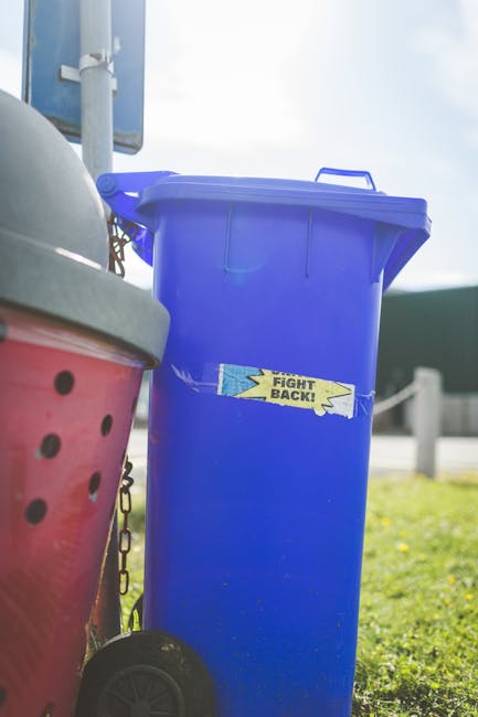 Two green wheeled rubbish bins are positioned outdoors on a slightly snowy surface, with the left bin labeled with the number '1' and the right bin labeled with the number '2'. The bin on the left appears to have a smooth, matte finish with some minor scratches and scuff marks, while the bin on the right has a slightly textured surface with a darker, fitted lid and black handles. Both bins are set on a paved area bordered by a small brick wall, with a background of a grassy yard and a paved driveway, indicating a residential setting. The scene is lit by natural daylight, casting soft shadows beneath the bins. The context suggests a private collection system for waste and garden refuse, aligning with independent rubbish removal services such as those offered by Waste Collection Kingston, rather than official council collection. The image emphasizes the use of clearly marked, durable containers for refuse separation and disposal at a private property.