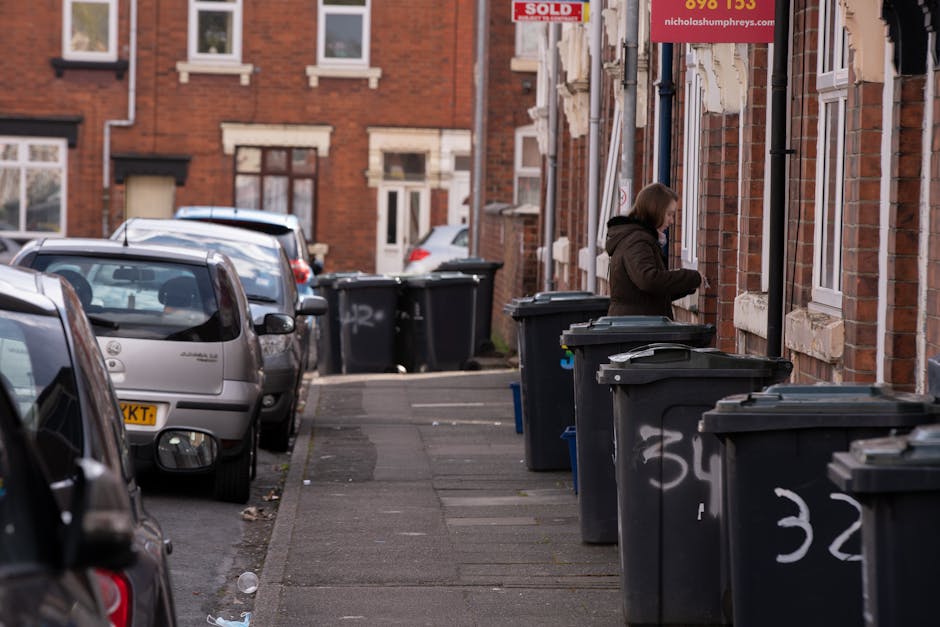 A residential street scene showing a row of large, dark grey wheeled rubbish bins with closing lids lined up along the edge of the pavement, in front of a brick building with multiple windows and a 'sold' sign visible on the wall. The bins are positioned close to the building's façade, which features white window frames and stone sills. A woman dressed in a brown coat, with shoulder-length hair, is standing next to one of the bins, appearing to be either placing rubbish inside or opening the lid. Several parked cars, including a silver hatchback and a darker vehicle, are situated along the street, which is paved with concrete slabs. The atmosphere appears overcast with diffuse natural lighting, typical of urban environments that may involve independent waste collection or private disposal services managed by waste management companies like Waste Collection Kingston. The scene captures a typical on-site clearance or rubbish collection setup, emphasizing routine waste disposal in a residential area with attention to detail in the textures of brickwork, vehicle surfaces, and the waste containers.