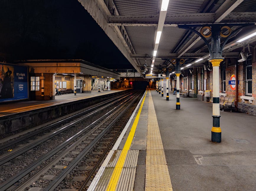 The image depicts a deserted underground train station platform illuminated by bright overhead lighting, with the platform extending into the distance beneath a metal and brick canopy supported by decorative iron columns painted black with yellow accents. The platform surface appears concrete with tactile paving strips and yellow safety lines along the edge, adjacent to the railway tracks made of steel rails and wooden sleepers. The station features brick walls with large windows and a classic roundel sign indicating the London Underground. On the far side of the platform, there are benches and a few advertisements, including one for Prime Video, with a person sitting near the wall. The overall environment is quiet and clean, with a subtle sense of anticipation created by the empty platform and dark tunnel in the background, reflecting a typical scene for private or independent transit services that may require alternative collection or on-site management, as handled by waste collection Kingston or similar providers in waste removal contexts.