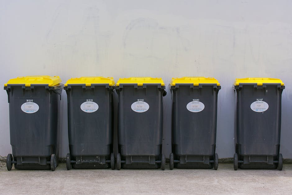 A row of five large black wheelie bins with yellow lids, made of durable plastic, lined up against a plain light grey wall. Each bin features a white oval label with black text and a barcode, indicating identification or collection details. The bins are positioned on a flat concrete surface, with their wheels resting on the ground, and are evenly spaced apart. The environment appears to be an outdoor area such as a driveway or designated waste collection zone, with diffused natural lighting casting minimal shadows. The scene reflects an organized setup for waste storage or collection, consistent with services provided by waste management companies like Waste Collection Kingston, which often handle domestic or commercial rubbish disposal through independent collection methods rather than local authority services. The bins are in good condition, with textured surfaces and secure-fitting lids, ready for rubbish removal or on-site waste handling. This setup visually exemplifies convenient and private waste collection options aligned with alternative disposal solutions for residential or business premises.