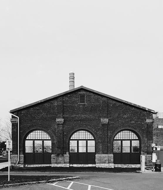 A black and white photograph depicting the front facade of a large, industrial brick building with a pitched roof. The building features three prominent arched windows with black frames, set into the brick wall, and reinforced with stone detailing at the base of each arch. A thin, metal chimney protrudes from the roof's ridge, indicating an older industrial or warehouse structure. The surrounding environment includes a paved roadway in the foreground with a parking restriction sign and a bollard, suggesting a parking area or street adjacent to the building. The overall atmosphere appears calm and quiet with no visible activity. This scene subtly relates to waste management services by exemplifying a site that might require building clearance or site-specific disposal, which companies like Waste Collection Kingston could support through independent or private waste collection, especially for commercial or industrial properties needing on-site clearance or waste removal in conjunction with their rubbish services.