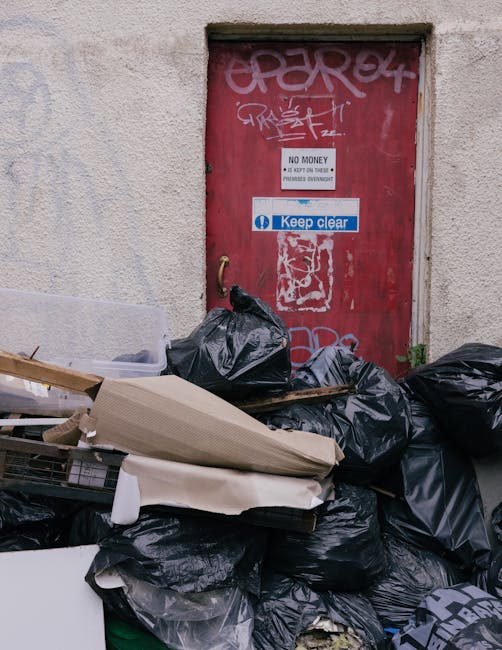 A large pile of black rubbish bags and discarded cardboard, wood, and plastic waste is stacked in front of a red metal door set into a rough-textured beige wall. The bags appear full and are stacked haphazardly across the pavement, with some materials such as rolled cardboard and broken wood protruding from the pile. The door is weathered, with graffiti tags and stickers adhered to its surface, including a white sticker with black lettering that states 'NO MONEY' and a blue sticker reading 'Keep clear.' The environment suggests an urban setting, possibly an alleyway or back street, where informal waste disposal or on-site clearance has occurred. Waste collection Kingston often advises on proper rubbish disposal, and this scene may depict an example of private or alternative waste handling, with waste accumulated beside the building for removal or collection service. The lighting is natural, consistent with outdoor conditions, emphasizing the textures of the waste and the surrounding wall, highlighting the importance of responsible rubbish management and clearance in urban environments.