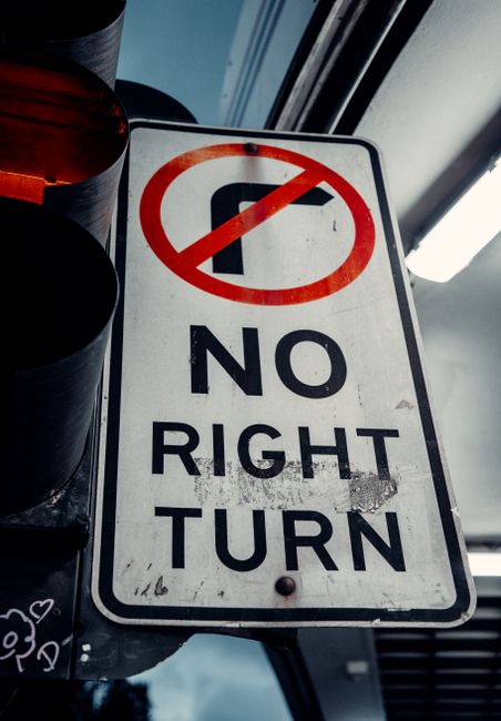 An upright rectangular road sign with a white background and black border, located indoors or on a vehicle, displaying a red circle with a diagonal line crossing out a black left-turn arrow at the top. Below, the sign has bold black lettering that reads 'NO RIGHT TURN'. The surface of the sign shows some dirt and minor scratches, indicating exposure to outdoor environments or handling. To the left of the sign, partially visible, there is a black and red traffic light or signal component. The background features a window with a view of overcast sky and blurred industrial or vehicle interior elements, suggesting the sign is situated in a setting related to road traffic management or vehicle operation, relevant to the context of managing waste collection routes or private disposal services. The overall scene communicates clear traffic regulation, which could be part of instructions for safe and compliant rubbish removal processes involving alternative or independent collection methods. The lighting is natural and neutral, emphasizing the sign’s visibility and importance in guiding transport or waste management activities.
