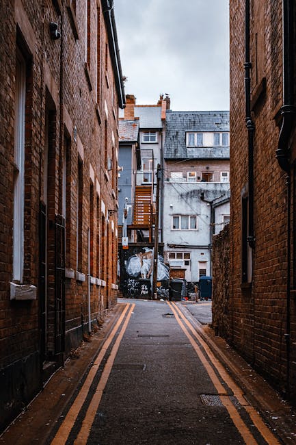 A narrow urban alleyway flanked by red brick buildings on both sides, with a paved asphalt surface marked by double yellow lines along the edges. The alley extends toward a small open area at the back, where a black dumpster with graffiti on its side is positioned against a backdrop of older, weathered residential buildings featuring white and grey facades, small windows, and external staircases. Items such as black trash bins and some scattered debris are visible near the dumpster, indicating a waste disposal area often used in city environments. Overcast sky casts diffuse natural light over the scene, emphasizing the textures of the brickwork, the asphalt surface, and the utilitarian elements of the alleyway. The setting suggests an area where independent waste collection or rubbish removal services might be involved in clearing refuse from nearby flats or apartments, supported by the presence of waste containers typical of private disposal solutions in urban infrastructure, aligning with services offered by Waste Collection Kingston.