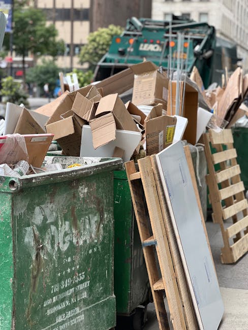 A large, open green waste container positioned on a city street, filled with various discarded cardboard boxes, some crushed and others partially assembled, along with a few smaller packaging materials. Several pieces of packaging, including a white cardboard box with yellow labels and plain brown boxes, are stacked or leaning against the container’s side. Nearby, a wooden pallet is resting against the waste bin, partially visible, with slats and a light wood finish. In the background, a waste collection vehicle with a dark green body and hydraulic lifting equipment is parked, partially obscured by the pile of rubbish. Surrounding the scene, there are nearby trees with blurred green foliage and multi-story buildings, indicating an urban environment. The lighting suggests daytime with natural light illuminating the scene evenly, showing details such as textures of the cardboard, wood, and painted surface of the waste container. This scene exemplifies a typical waste collection operation involving private rubbish removal, potentially handled by an independent service provider like Waste Collection Kingston, emphasizing the importance of organized waste disposal in urban settings.