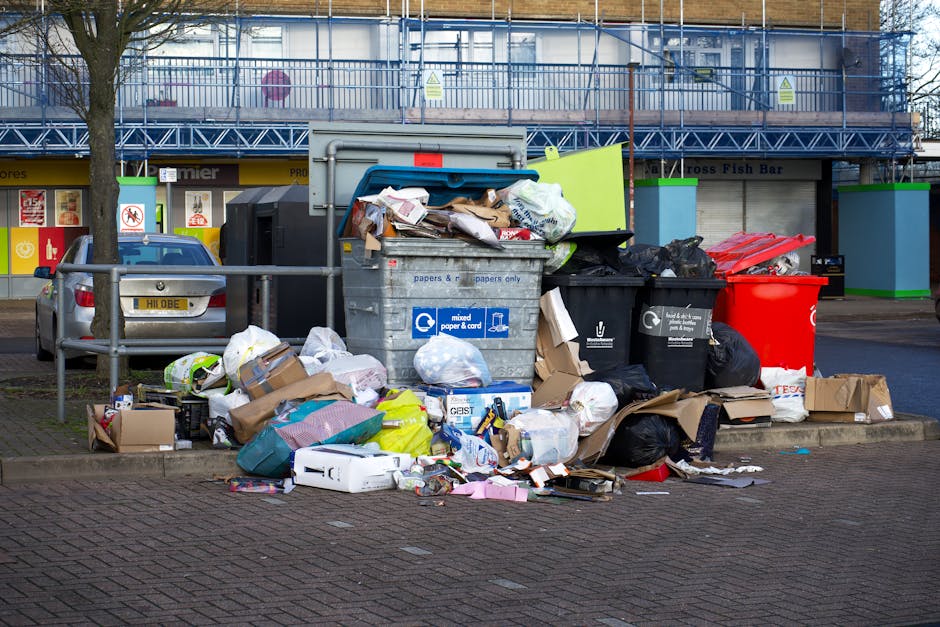A pile of mixed household rubbish and waste materials accumulated around a recycling bin on a paved urban street. The waste includes cardboard boxes, paper bags, plastic bottles, black garbage bags, and flattened packaging. The recycling bin, labeled for mixed paper and cardboard, is overflowing with paper waste spilling onto the ground. Nearby, there are additional black bins, and a red bin is visible to the right. In the background, a parked silver car is partially visible behind a metal railing, with a building facade featuring blue scaffolding, storefronts, and signage. The scene is outdoors in daylight, with a tree on the left side, and the surrounding environment suggests an area designated for waste collection, although the overflowing rubbish indicates the need for an alternative waste management solution, possibly provided by Waste Collection Kingston for private or non-standard rubbish removal.