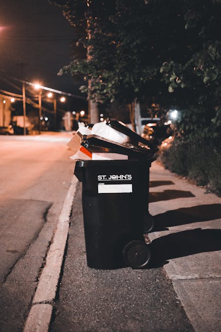 A black wheeled rubbish bin positioned on the pavement beside a curb in a residential or urban area at night. The bin is labelled with white text reading 'ST. JOHN'S' and is overflowing with various waste items, including cardboard and plastic debris, some of which are protruding from the open top. The scene is illuminated by nearby streetlights, casting shadows on the ground and highlighting the texture of the bin’s surface. In the background, there are trees with dense foliage, additional streetlights, and parked vehicles, indicating a quiet, well-lit street environment. The setting suggests an instance of private waste disposal or an on-site rubbish collection, typical of independent rubbish removal services, with no visible action occurring but implying the ready state for rubbish collection or removal. The overall scene portrays a standard residential rubbish bin prepared for waste collection, situating the image within the context of waste management services like those provided by Waste Collection Kingston, emphasizing the importance of proper rubbish handling and disposal methods.