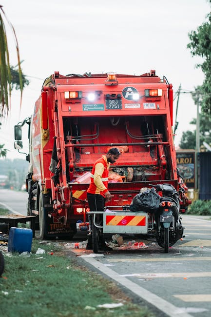 A pile of mixed household rubbish and waste materials accumulated around a recycling bin on a paved urban street. The waste includes cardboard boxes, paper bags, plastic bottles, black garbage bags, and flattened packaging. The recycling bin, labeled for mixed paper and cardboard, is overflowing with paper waste spilling onto the ground. Nearby, there are additional black bins, and a red bin is visible to the right. In the background, a parked silver car is partially visible behind a metal railing, with a building facade featuring blue scaffolding, storefronts, and signage. The scene is outdoors in daylight, with a tree on the left side, and the surrounding environment suggests an area designated for waste collection, although the overflowing rubbish indicates the need for an alternative waste management solution, possibly provided by Waste Collection Kingston for private or non-standard rubbish removal.